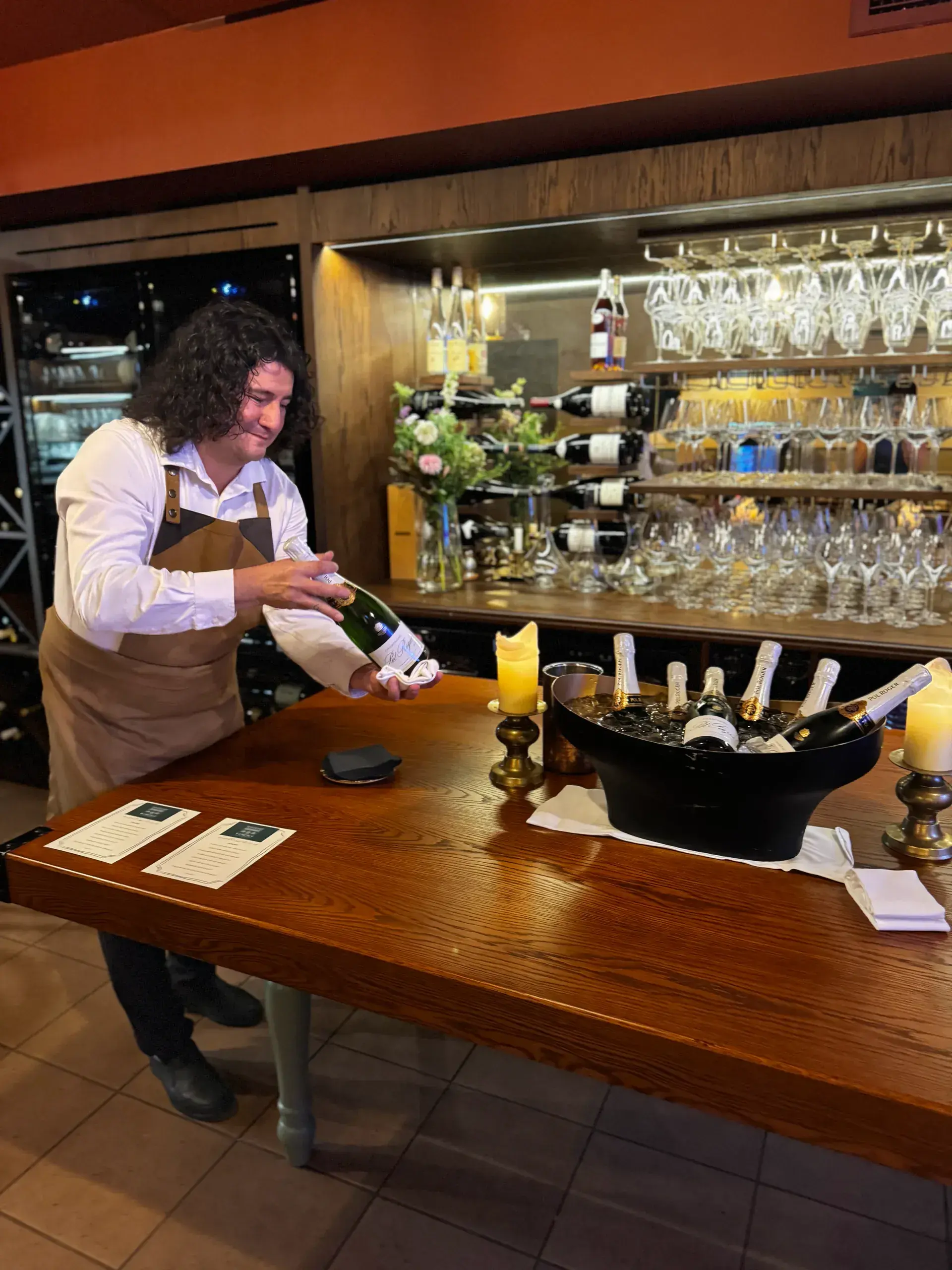 A host presents a bottle of Pol Roger on a napkin at the tasting table, in front of the lit back bar