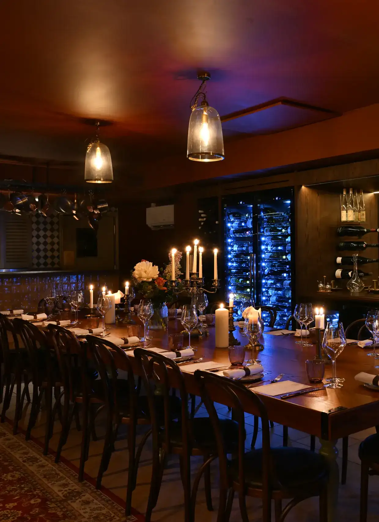 The long table dressed for dinner under glass pendant lamps, with lit candles, a candelabra and flowers, and the lit wine fridges beyond