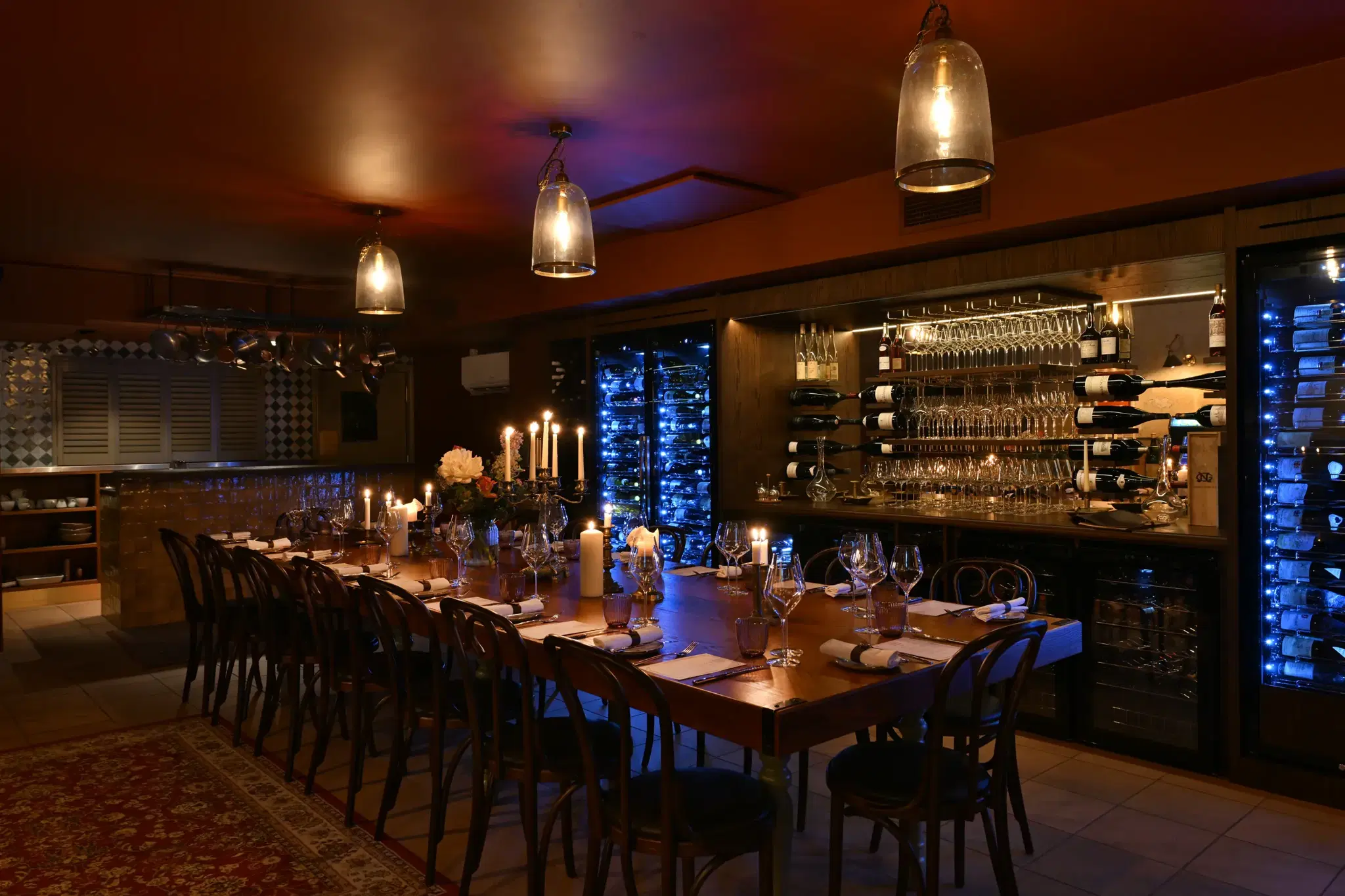 The long table dressed for dinner, with lit candles, a candelabra and flowers, beside the lit wine fridges and the bar