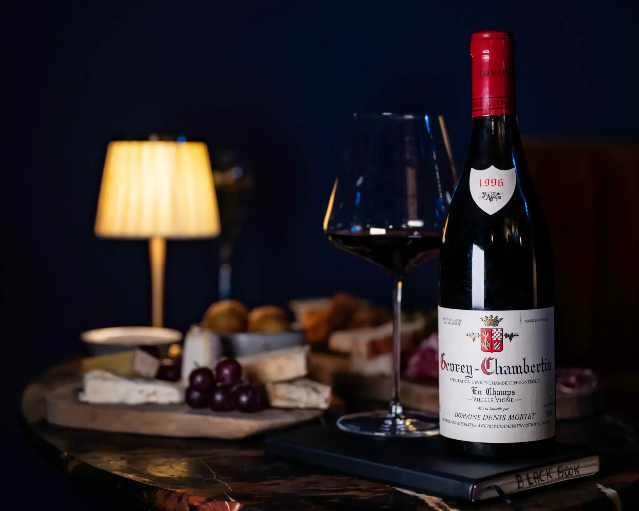 A 1996 Gevrey-Chambertin beside a glass of red wine, lit by a table lamp against a deep blue wall