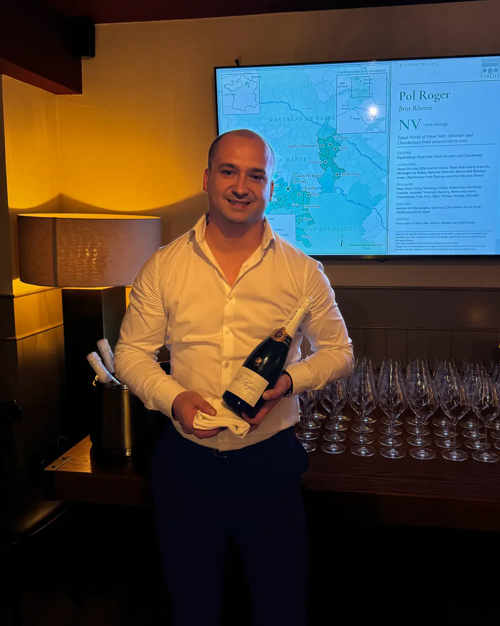 A host presents a bottle of Pol Roger in front of a screen showing a map of Champagne and the tasting notes, with glasses set out for a tasting