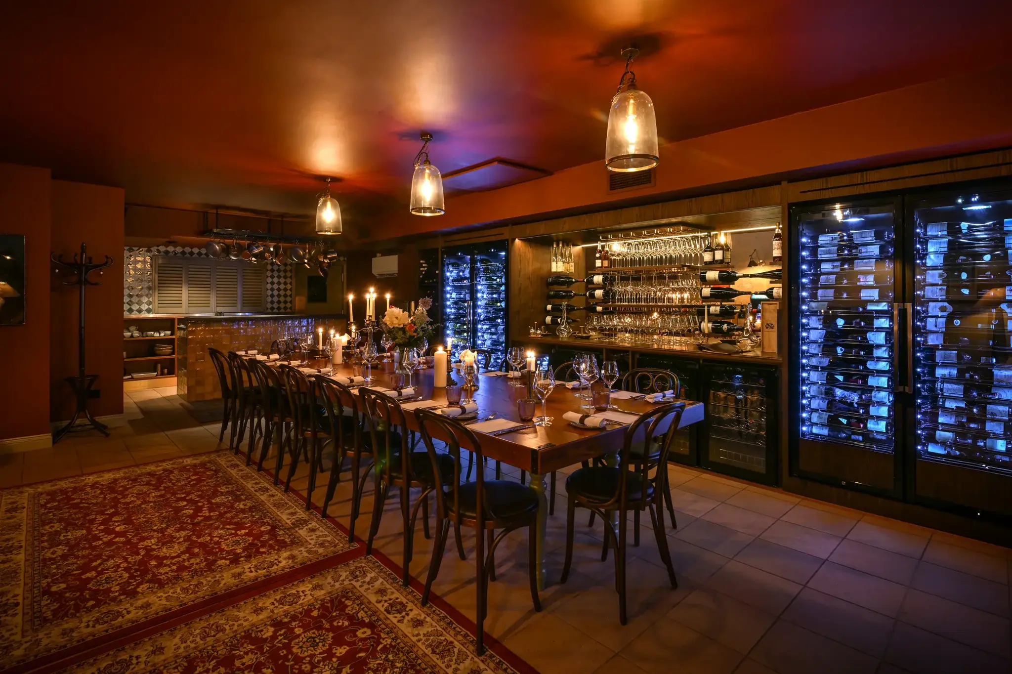 The private dining room looking towards the kitchen: the long candlelit table beside Persian rugs, the lit wine fridges and the back-lit glassware of the bar